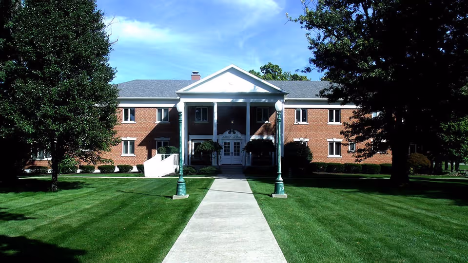 Front exterior view of Judson Palmer Home, a two-story brick building with white columns at the entrance, surrounded by green grass and trees under a blue sky.