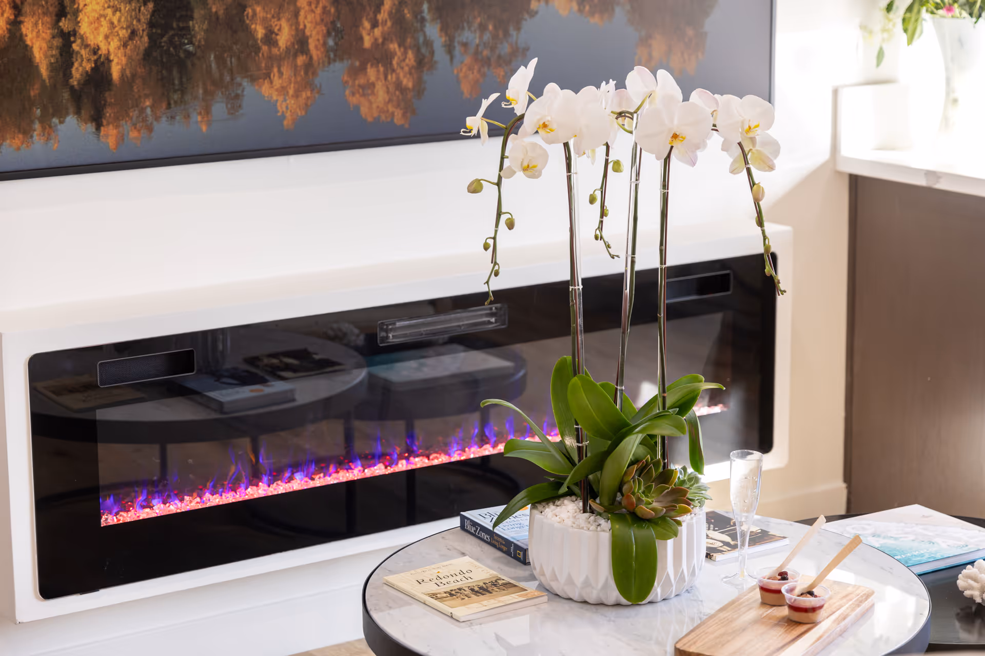 A modern electric fireplace with blue and orange flames beneath a large framed picture. In front of the fireplace is a round marble table with a white orchid plant in a white pot, several books, a glass of champagne, and two small dessert cups with wooden spoons.
