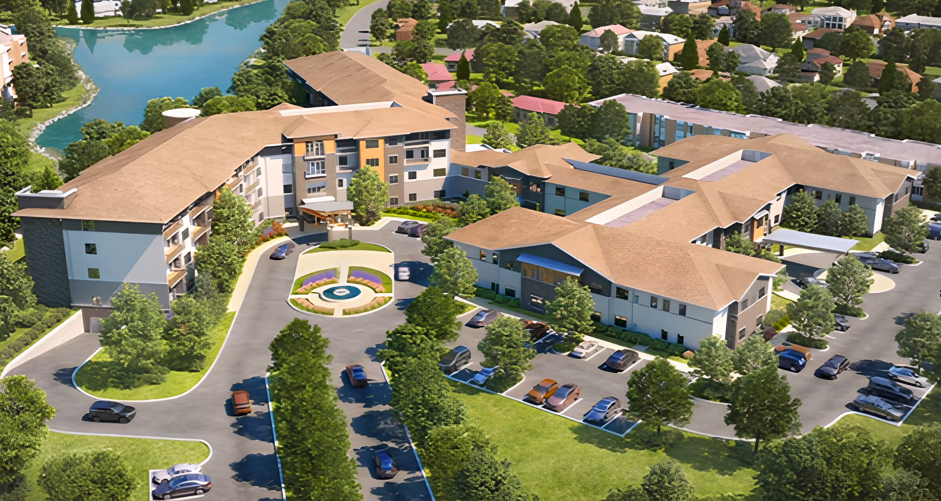 Aerial view of Friendship Village Retirement Community showing multiple connected buildings with tan roofs surrounded by trees and greenery. There is a circular driveway with a central landscaped area and fountain, several parking lots with cars, and a body of water visible in the background.