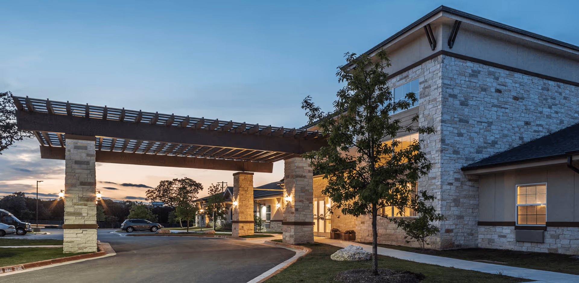 Exterior view of a senior living facility at dusk with a covered entrance supported by stone pillars, a tree in the foreground, and cars parked nearby under a clear sky.