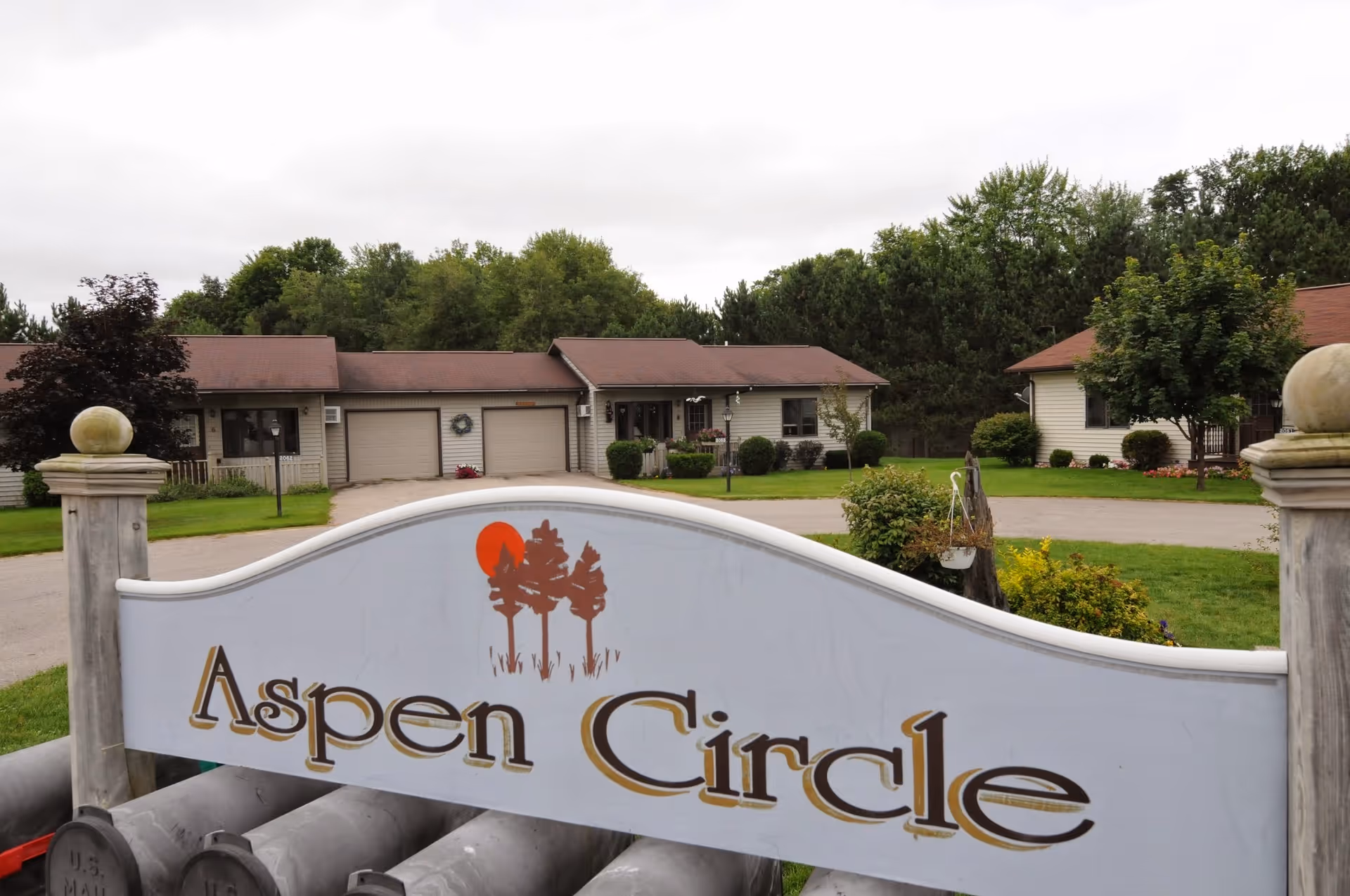 White curved sign reading "Aspen Circle" in front of single-story residential buildings, lawn, and trees.