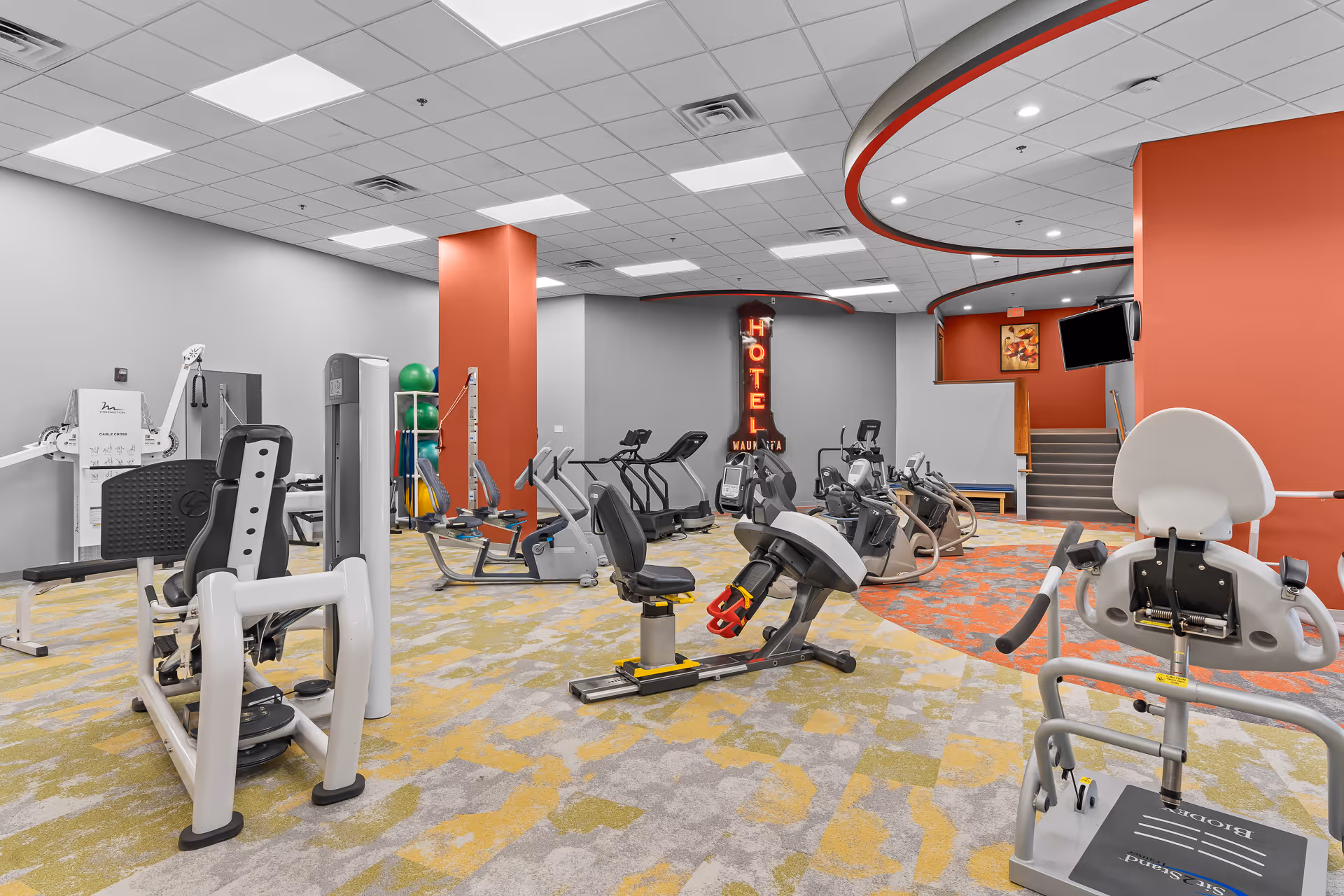 A spacious fitness room with various exercise equipment including stationary bikes, weight machines, and resistance training devices. The room has a patterned carpet in shades of yellow, gray, and orange, with orange accent walls and a ceiling with recessed lighting. A neon sign reading 'HOTEL WAUKAFA' is visible on the back wall near a staircase and a mounted TV.