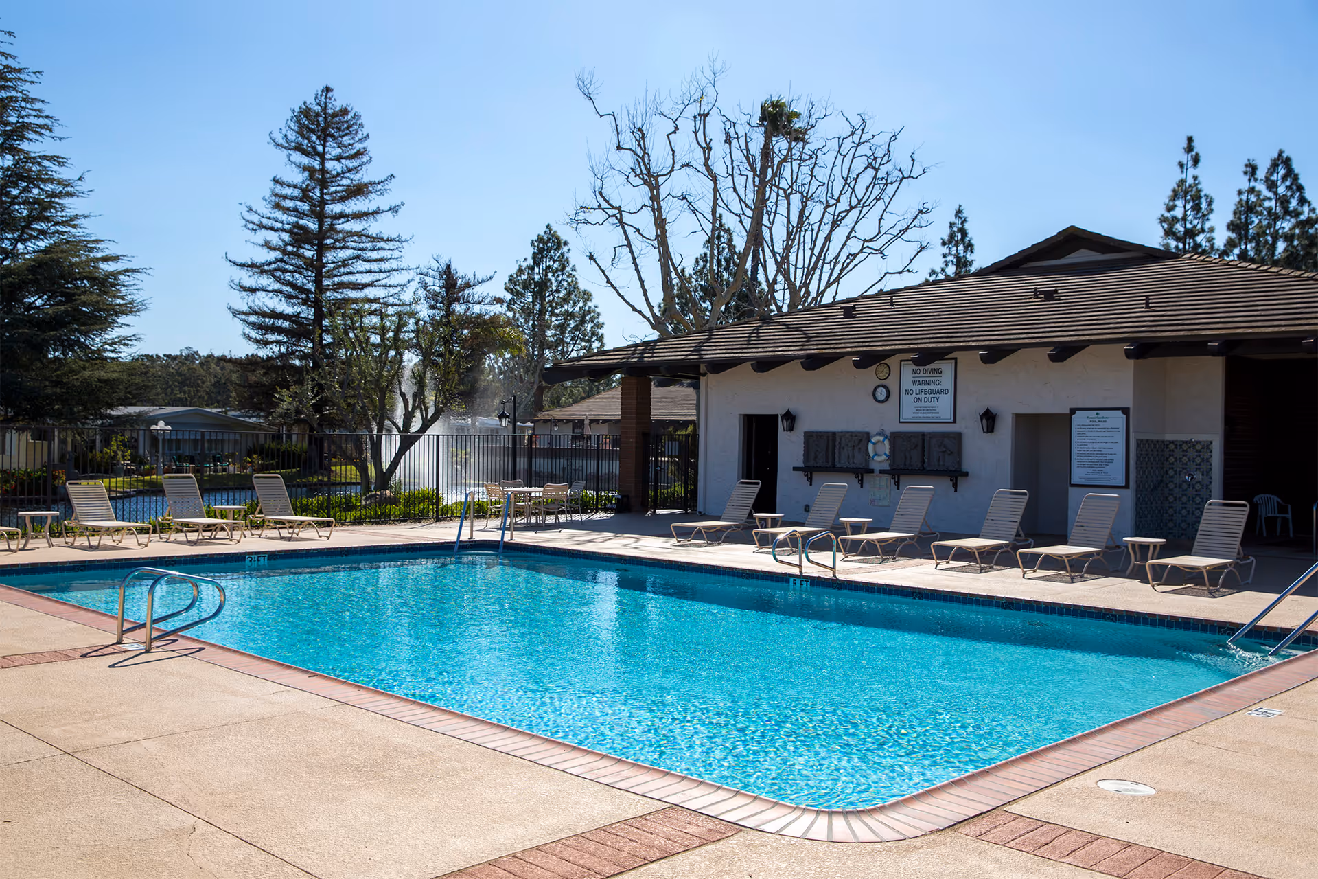 Outdoor swimming pool with clear blue water surrounded by beige concrete deck and several lounge chairs. In the background, there is a building with a tiled roof and signs on the wall, along with trees and a fountain visible behind a black metal fence.