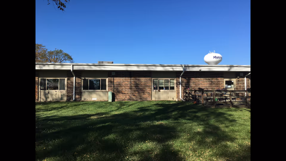 Single-story brick building exterior with windows, a small wooden porch, and a grassy lawn under a clear blue sky.