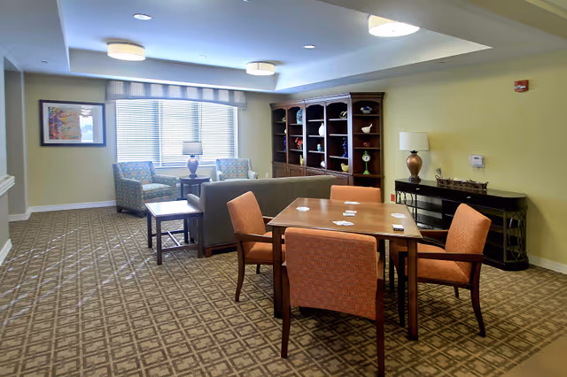 A well-lit common area in a senior living facility featuring a wooden table with four orange upholstered chairs, a gray sofa, two patterned armchairs near a window with blinds, a wooden bookshelf with decorative items, and two table lamps on side tables against beige walls and patterned carpet.
