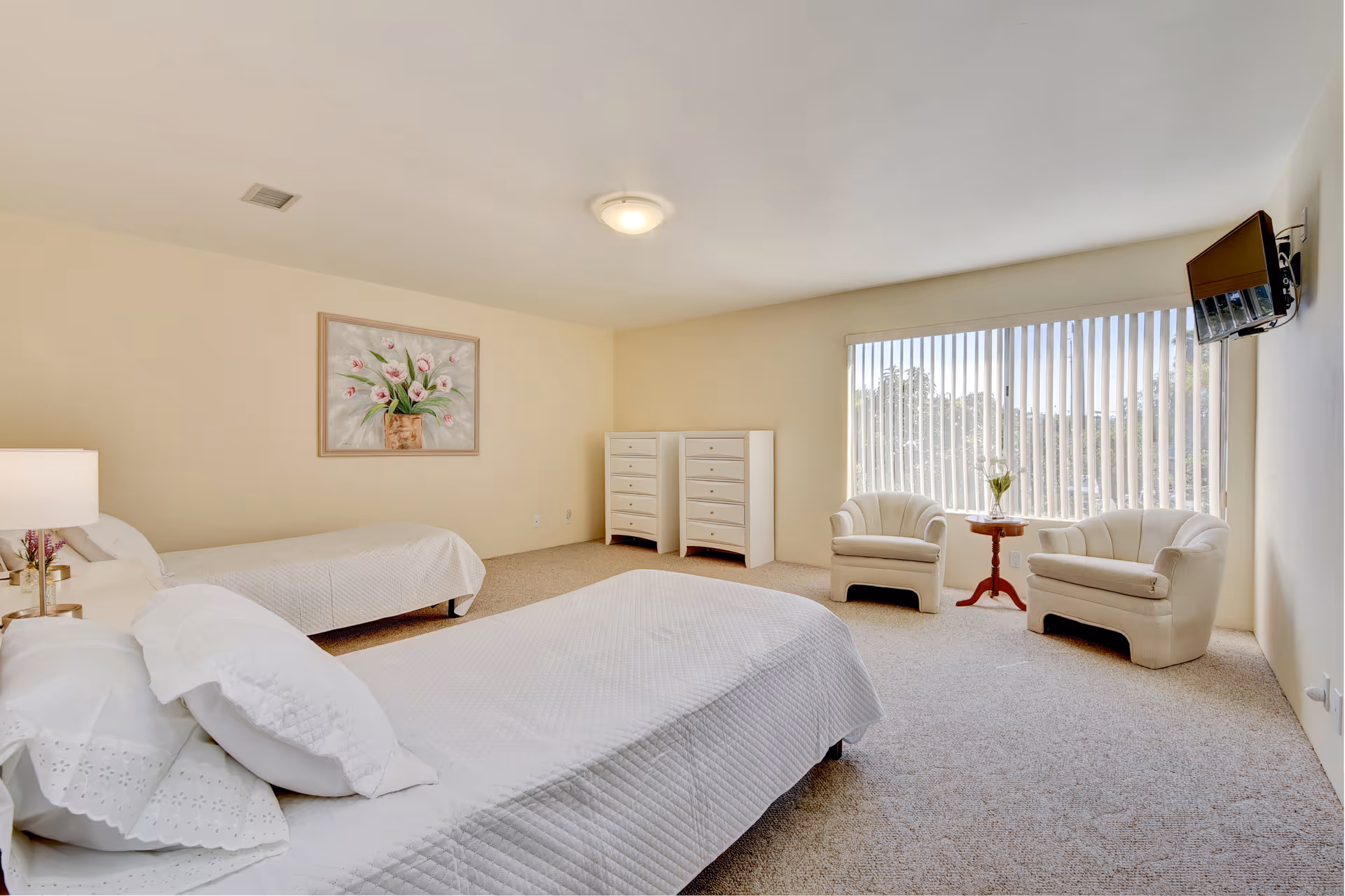 A spacious bedroom with two beds covered in white bedding, a nightstand with a lamp, a painting of flowers on the wall, two white dressers, two white armchairs with a small round wooden table between them, and a wall-mounted TV near a large window with vertical blinds.