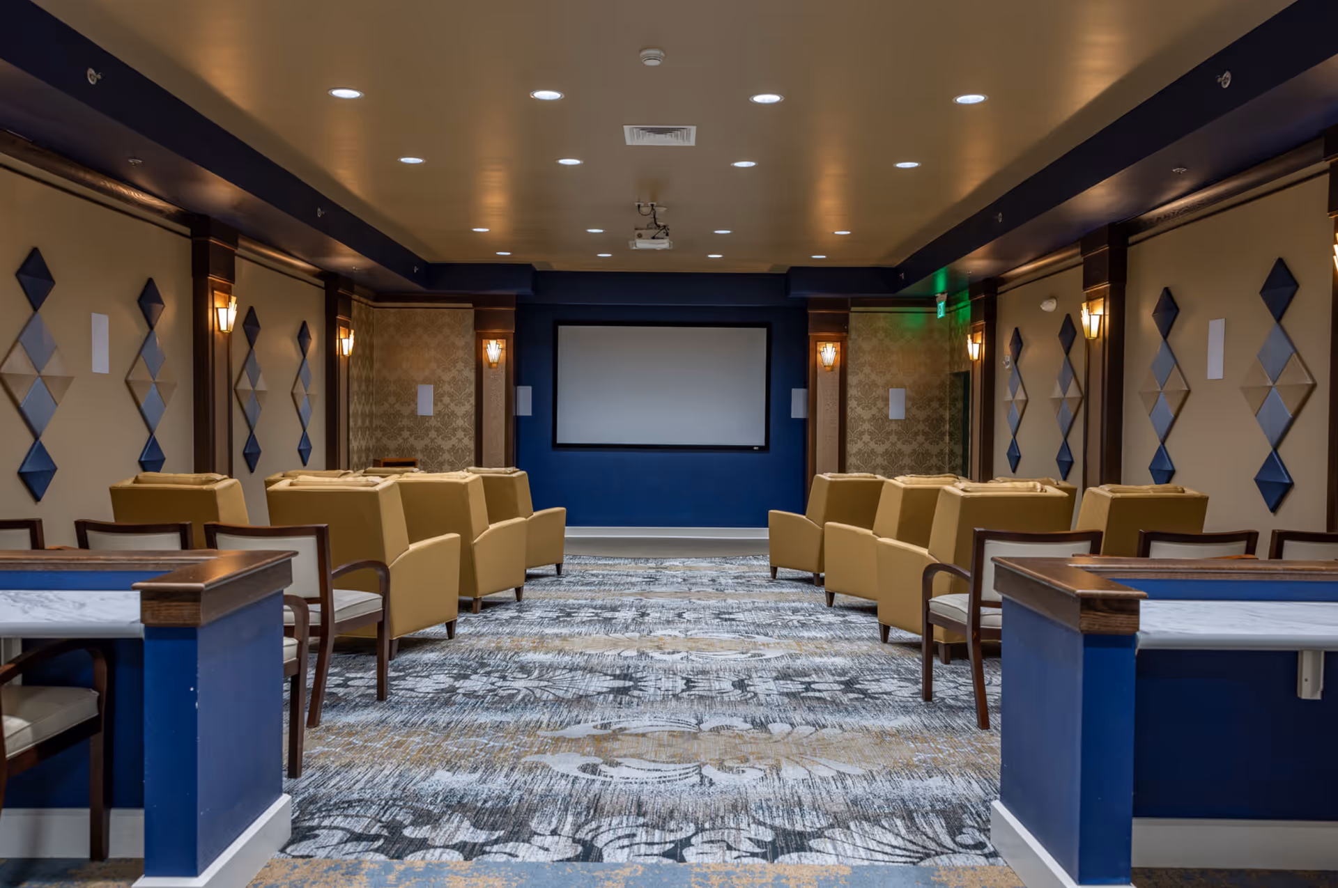 A cozy theater room with rows of beige armchairs facing a large blank projection screen on a dark blue wall. The room features patterned carpet, wall sconces, and decorative diamond-shaped wall panels in shades of blue and beige.