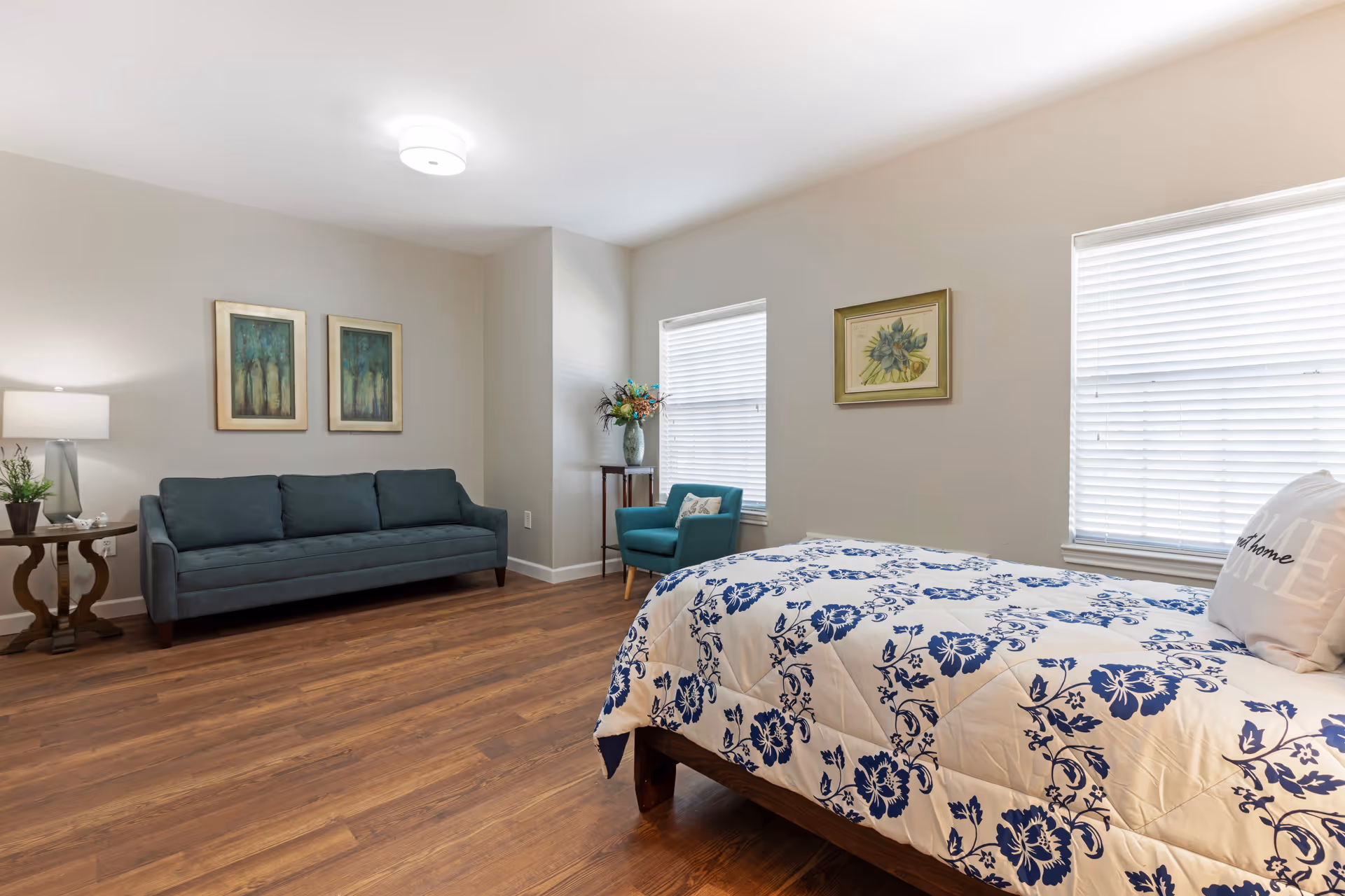 A senior living bedroom with a bed covered in a white and blue floral comforter, a teal armchair near two windows with blinds, a dark teal sofa against a wall with two framed pictures above it, a wooden side table with a lamp and a small plant, and wooden flooring.