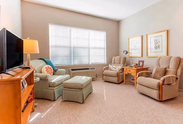 A cozy living room with a large window covered by white blinds. The room features a green armchair with a matching ottoman, two beige recliners with pillows, a wooden side table with a lamp and framed photos, and a wooden TV stand with a flat-screen television. The walls are light-colored and decorated with two framed botanical prints.