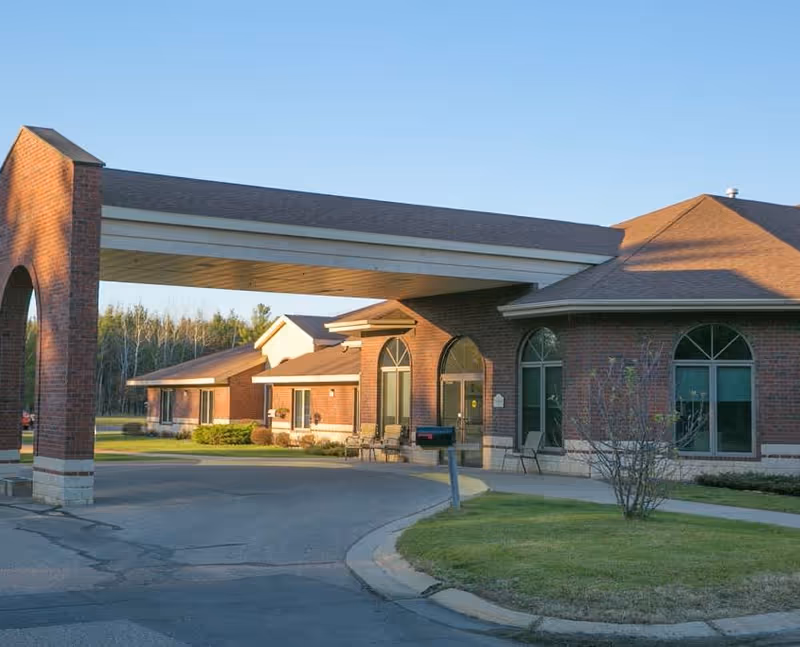 Exterior view of a senior living facility building with a covered entrance supported by brick pillars. The building has large arched windows and a driveway leading up to the entrance, surrounded by grass and some small bushes.