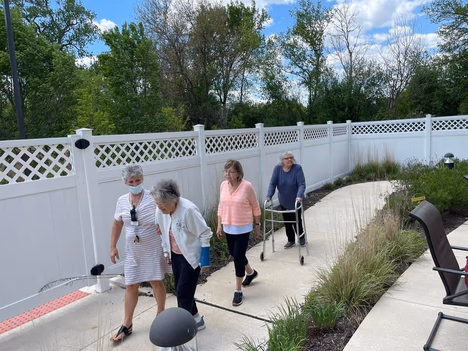 Four elderly women walking along a curved concrete pathway in an outdoor garden area enclosed by a white fence. One woman is using a walker, another is wearing a face mask, and the others are walking beside her. The garden has green plants and grass along the path, with trees and a partly cloudy sky in the background.