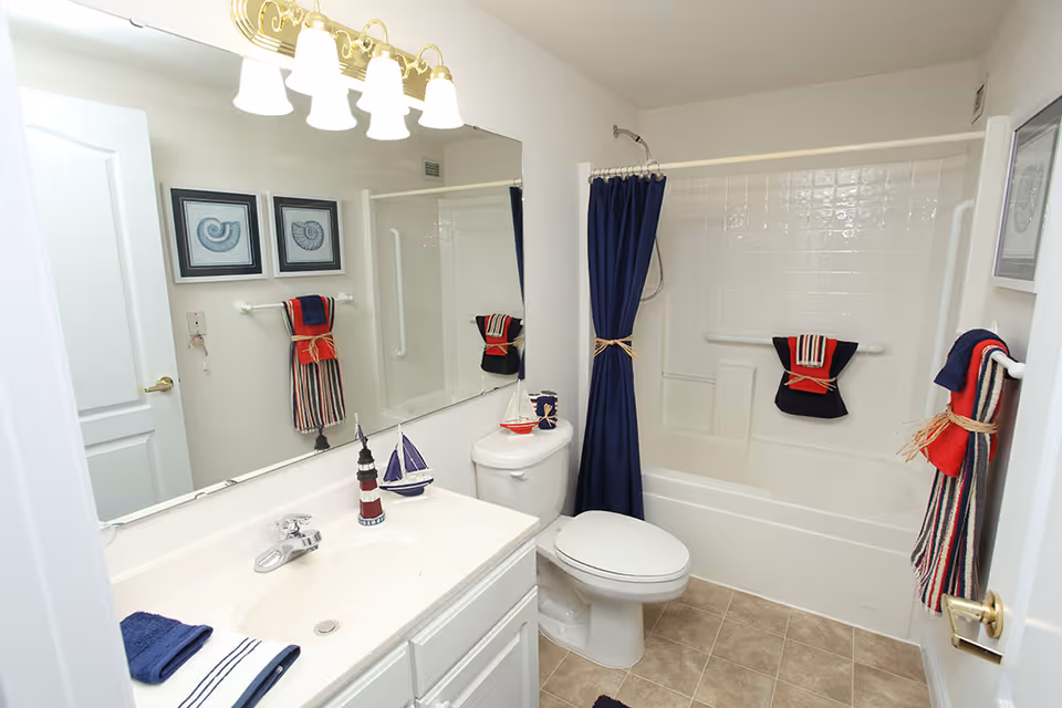 A clean and bright bathroom with a white sink and countertop, a toilet, and a bathtub with a shower. The shower has a navy blue curtain tied back with a rope. There are nautical-themed decorations including a small lighthouse and sailboat on the sink, and towels in red, white, and blue colors hanging on towel racks. Two framed pictures of seashells hang on the wall above the towel rack. The floor is tiled in a light brown color.