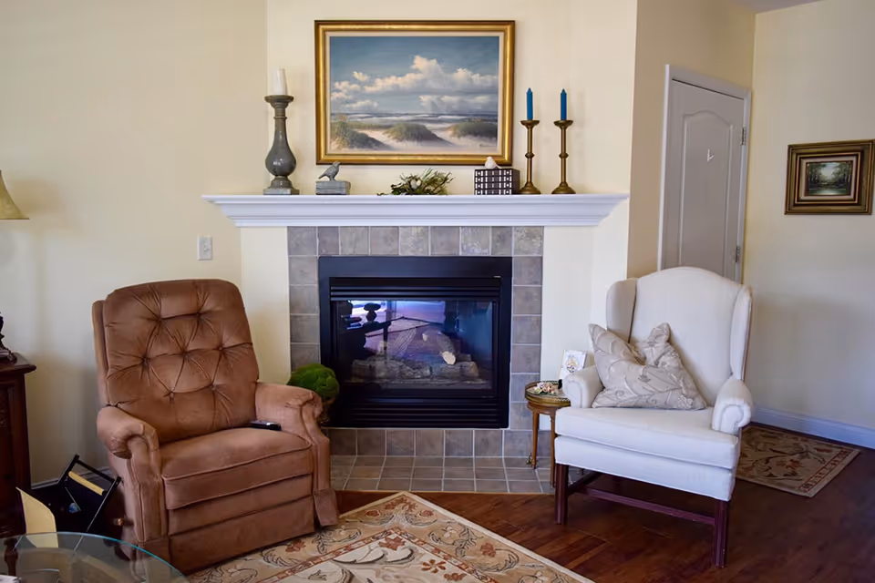 A cozy living room area featuring a brown cushioned recliner chair and a white armchair with decorative pillows, positioned on a patterned rug in front of a tiled fireplace. The fireplace mantel is adorned with candles, small decorative items, and a framed painting of a beach scene with clouds. The room has light-colored walls and wooden flooring.