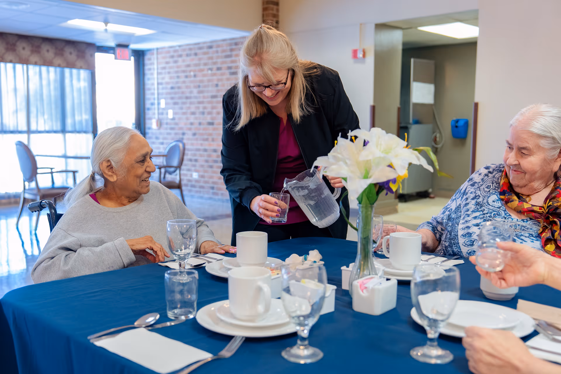 A woman is pouring water into a glass for an elderly woman seated at a table covered with a blue tablecloth. Two other elderly women are also seated around the table, which is set with white plates, cups, glasses, and silverware. A vase with white and purple flowers is in the center of the table. The setting appears to be a communal dining area with large windows and brick walls in the background.