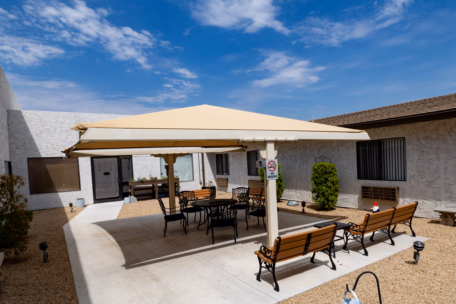 Outdoor courtyard area with a large beige canopy providing shade over a round table and several chairs. There are also wooden benches with black metal frames arranged around the shaded area. The courtyard is surrounded by a building with white textured walls and windows, and the ground is covered with gravel and concrete walkways. A clear blue sky with some clouds is visible above.