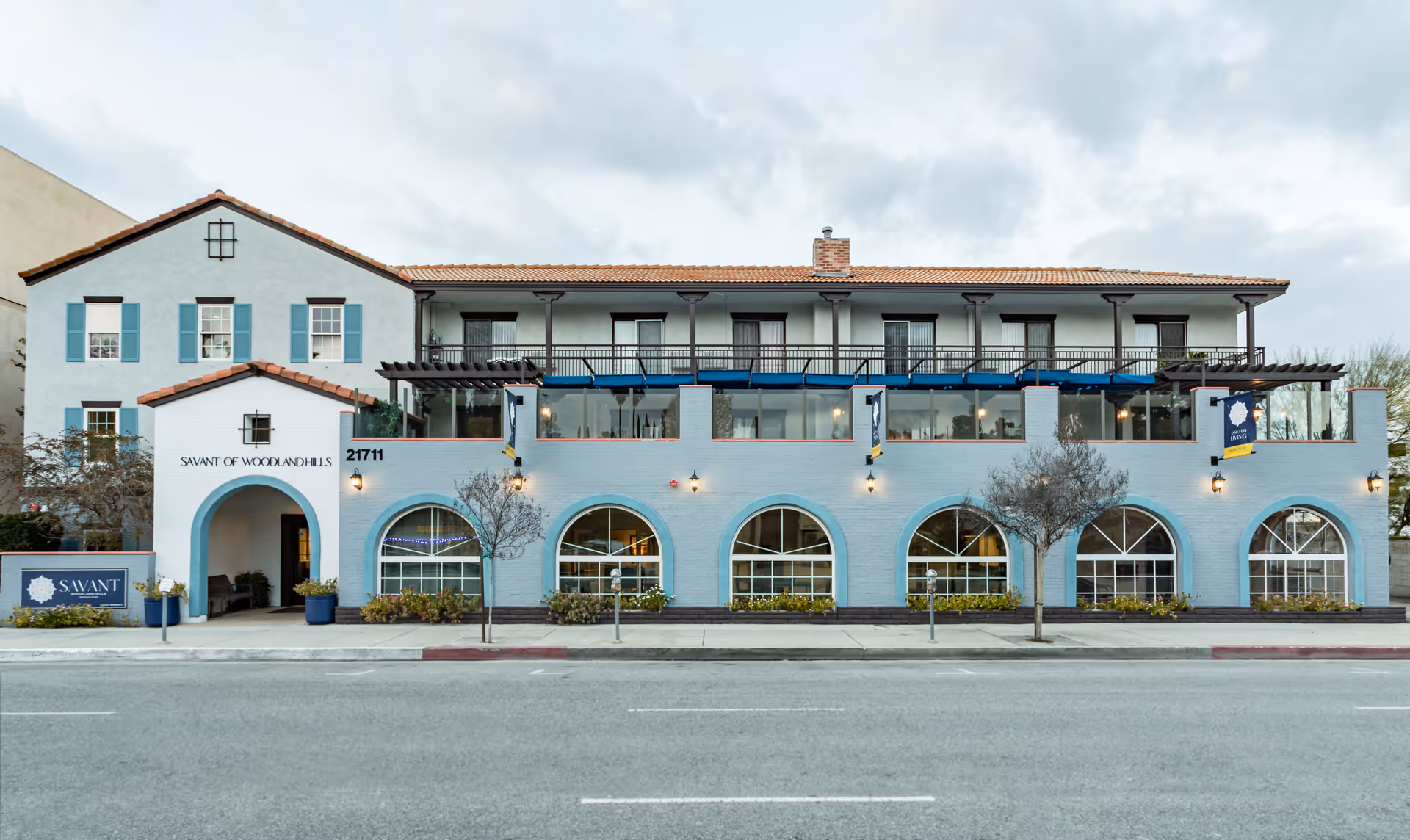 Front exterior view of Savant of Woodland Hills, a two-story building with light blue walls, arched windows on the ground floor, and a balcony with a pergola on the upper floor. The building has a tiled roof and a sign near the entrance displaying the facility's name.
