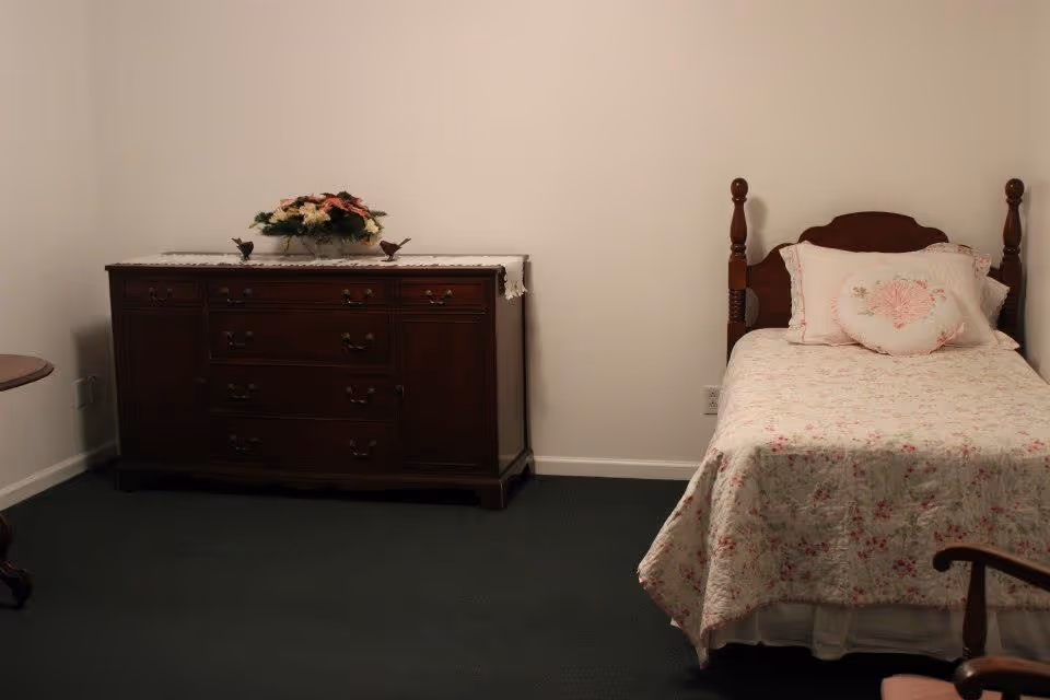 A simple bedroom with a single wooden bed covered with a floral bedspread and matching pillows. Next to the bed is a dark wooden dresser with multiple drawers, topped with a lace runner and a floral arrangement. The room has plain white walls and dark green carpet.