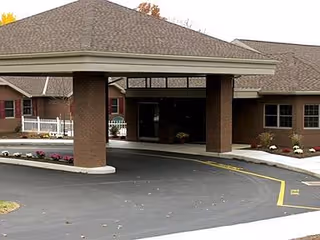 Entrance area of a senior living facility with a covered drop-off zone supported by brick pillars, surrounded by a paved driveway and landscaped with flowers and shrubs.