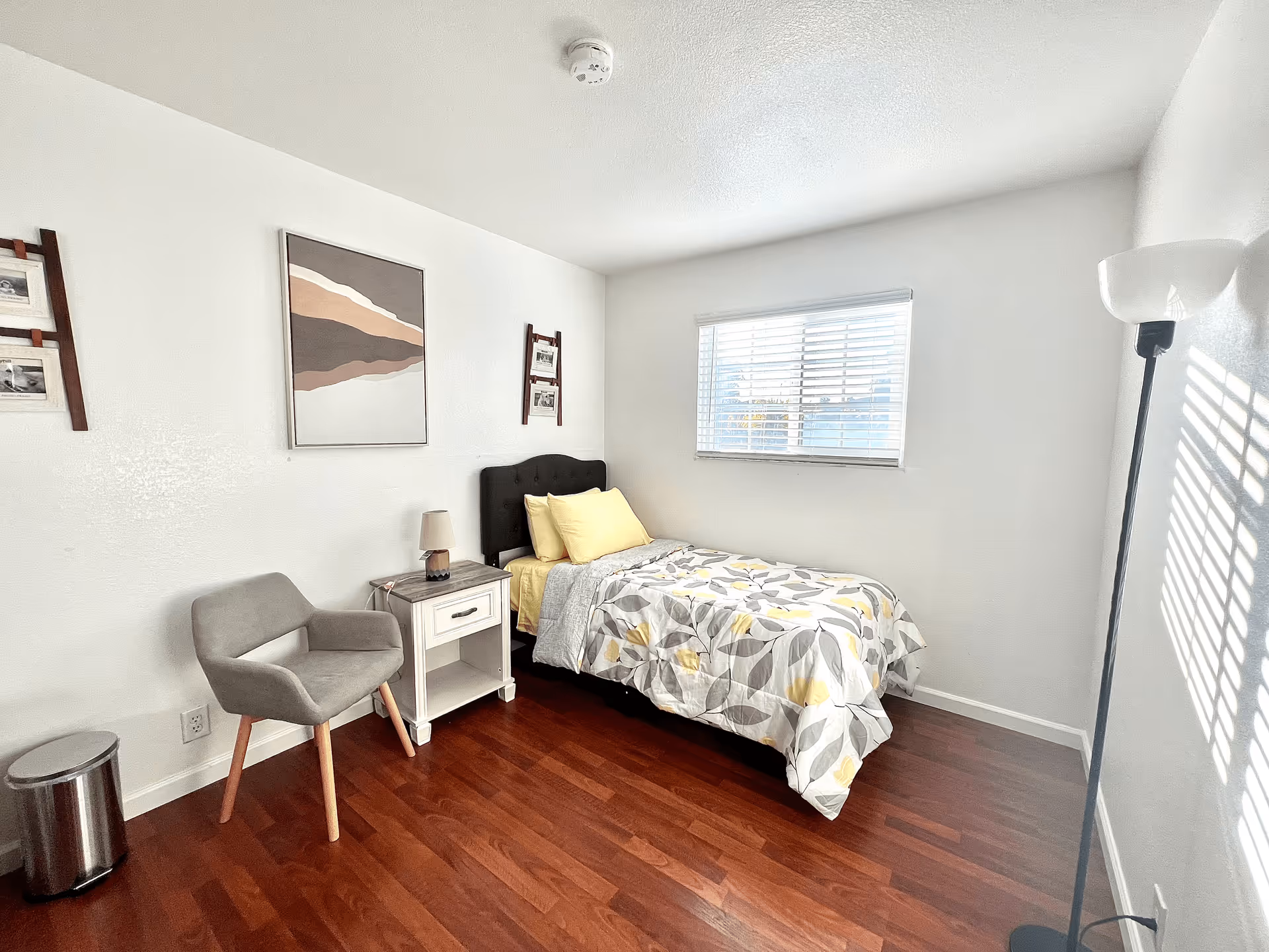 Small bright bedroom with a single bed, nightstand, gray chair, floor lamp, and a window letting in sunlight.