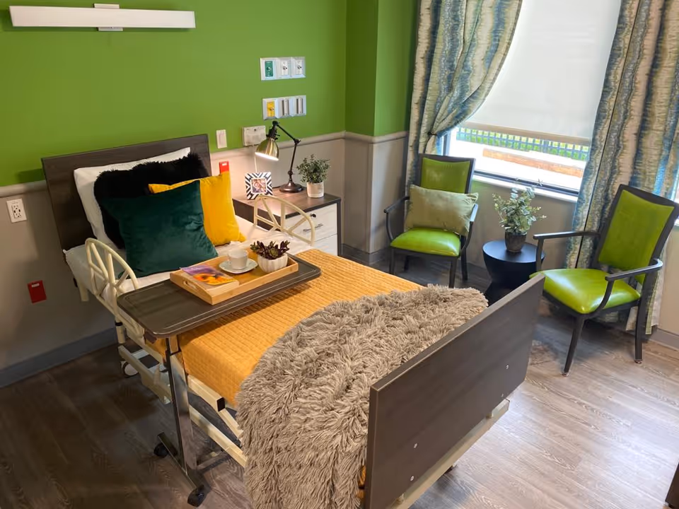 A neatly arranged nursing facility bedroom with a hospital bed covered in a yellow blanket and a gray furry throw. The bed has several pillows in black, green, and yellow. A tray with a cup, a small plant, and a book rests on the bed. Next to the bed is a nightstand with a lamp, a small plant, and a framed photo. Two green cushioned chairs and a small round black table with a plant are positioned near a window with blue and white curtains. The walls are painted green and gray, and the floor is wood.