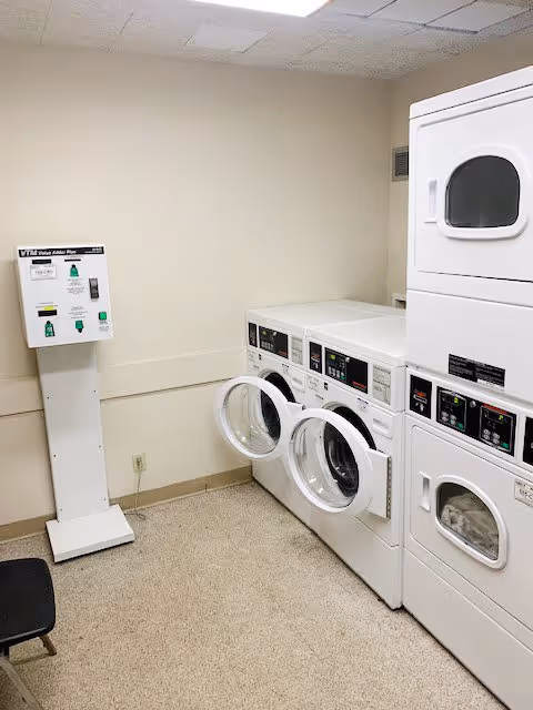Laundry room with two front-loading washing machines with open doors and two stacked dryers on the right side. A white vending machine or payment kiosk is positioned against the left wall. The room has beige walls and a speckled floor.