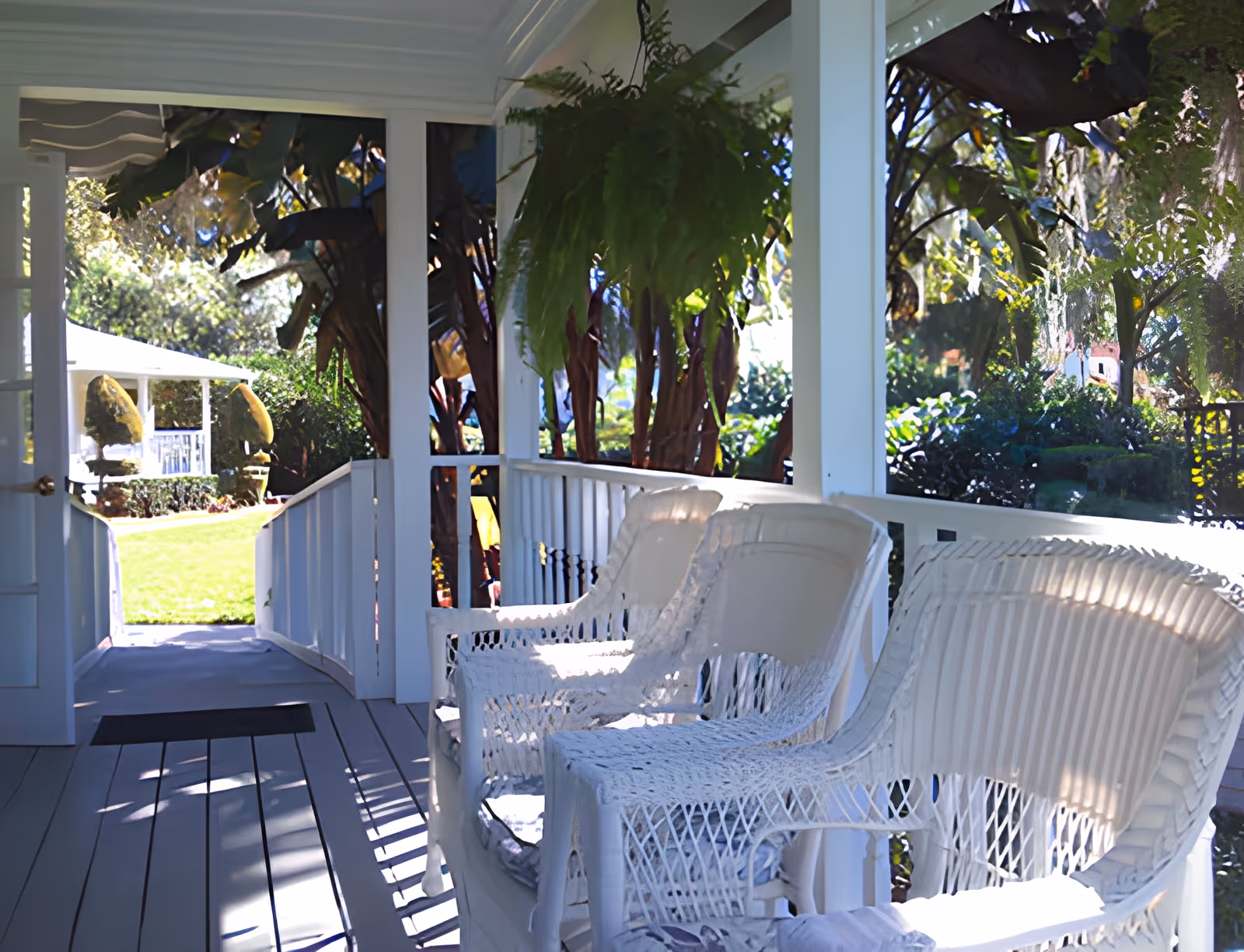 A bright and sunny porch area with white wicker chairs and a railing, overlooking a green garden with trees and shrubs. The porch has a wooden floor and white pillars, with a door open to the outside.