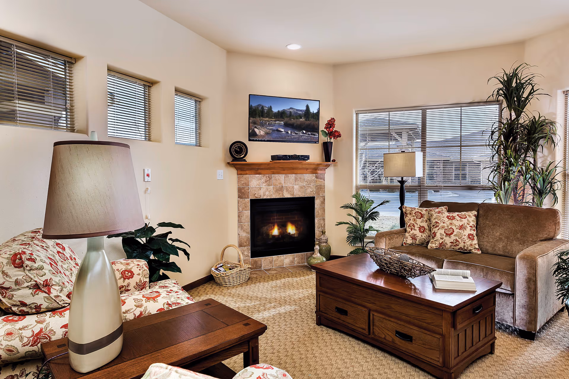 A cozy living room with a lit fireplace surrounded by beige tiled walls. There is a brown sofa with floral cushions, a floral armchair, a wooden coffee table with an open book and decorative bowl, and a side table with a lamp. Large windows with blinds let in natural light, and there are several green plants adding a touch of nature to the room. A flat-screen TV is mounted above the fireplace displaying a nature scene.