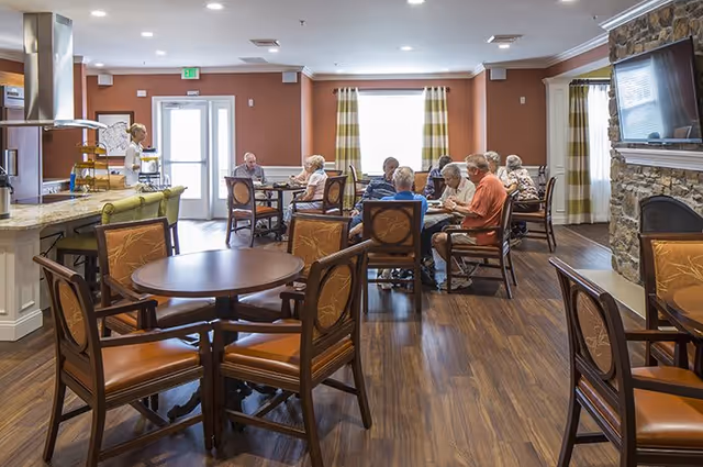 Several senior residents seated at tables in a communal dining and lounge area with a kitchen counter, stone fireplace, and wall-mounted TV.