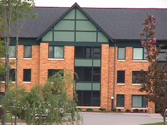 Exterior view of a three-story brick building with green trim and a dark roof, surrounded by trees and a paved area in front.