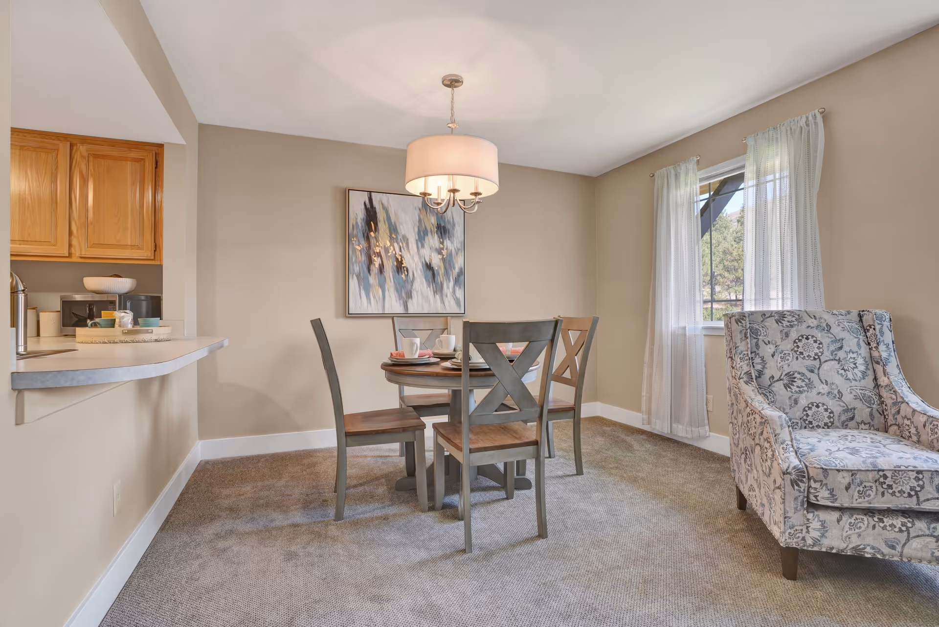 Small dining area with a round table and four chairs under a pendant light, a patterned armchair by a window, and a pass-through to the kitchen.
