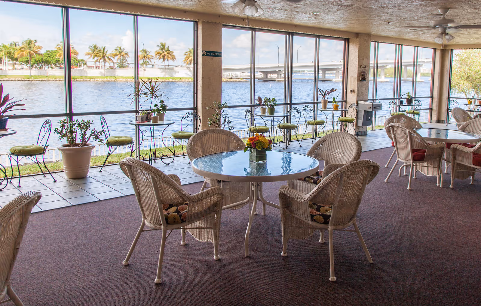 Indoor seating area with round glass-top tables and wicker chairs with cushions, overlooking a waterfront through large windows. Several small potted plants are placed on tables and along the window ledge. The space has carpeted flooring and ceiling fans.