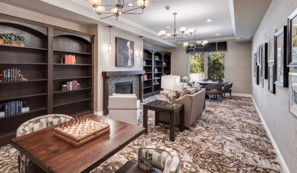 Cozy communal lounge with bookshelves, a fireplace, multiple seating areas, and a table with a chessboard.