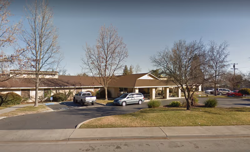 Exterior view of a single-story senior living facility building with a covered entrance, several parked cars, leafless trees, and a grassy area in front under a clear sky.