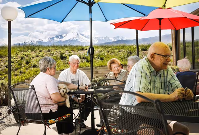 A group of elderly people sitting outdoors at metal tables with colorful umbrellas, enjoying a sunny day with a scenic mountain view in the background. One woman is holding a small dog on her lap.