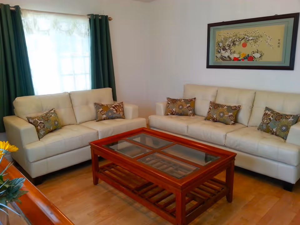 A living room with two cream-colored sofas adorned with patterned throw pillows. A wooden coffee table with a glass top is placed in the center on a wooden floor. There is a window with green curtains and sheer white drapes, and a framed artwork hangs on the wall above one of the sofas.
