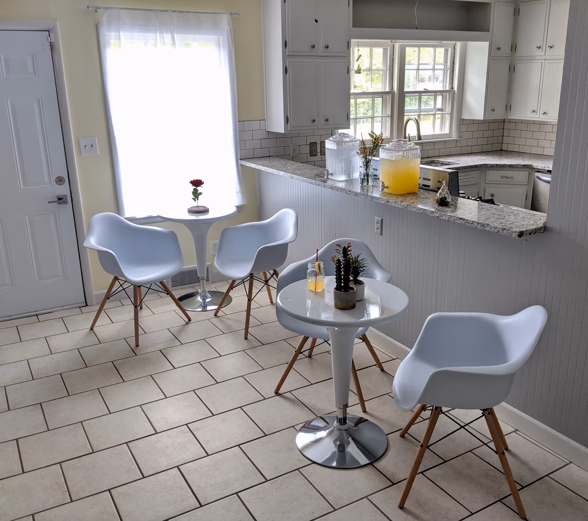 A bright kitchen dining area with two small white round tables, each surrounded by two modern white chairs with wooden legs. One table has a small plant and a drink with a straw, while the other has a single red rose in a vase. The kitchen counter has two large beverage dispensers, one with water and the other with a yellow drink, and a small plant. The room has tiled flooring, a window with sheer white curtains, and white cabinetry.