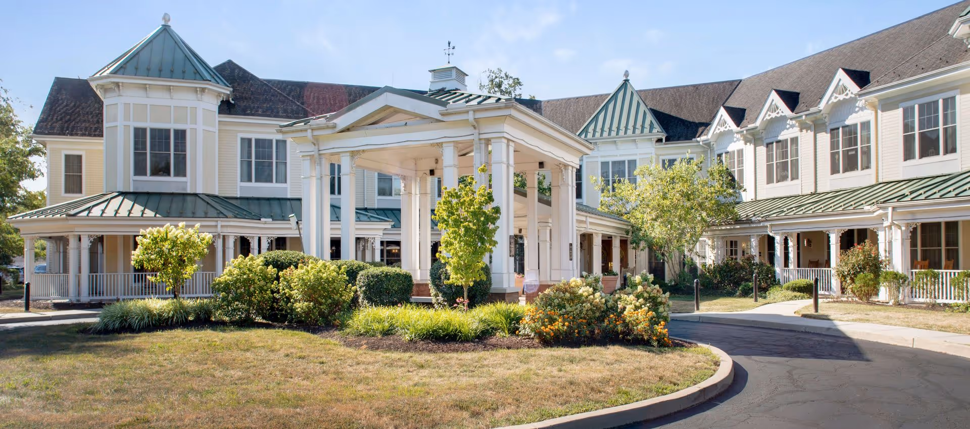 Exterior view of a senior living facility with a circular driveway, landscaped garden with bushes and flowers, and a covered entrance supported by white columns. The building has multiple windows, green metal roofs, and a light yellow facade under a clear blue sky.