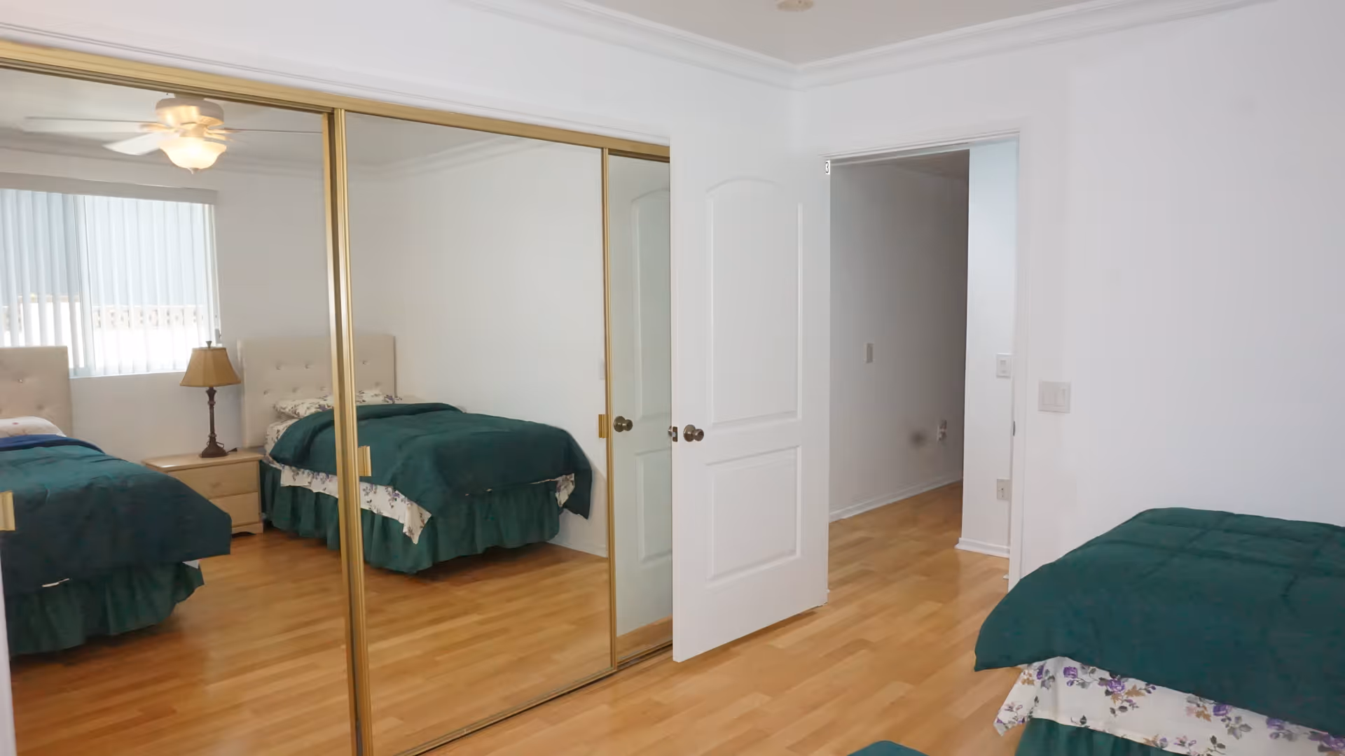 A bedroom with two twin beds covered in green blankets and floral sheets. There is a wooden nightstand with a lamp between the beds. A large mirrored sliding closet door reflects the beds and a ceiling fan with a light. The room has light wood flooring and white walls, with an open white door leading to a hallway.
