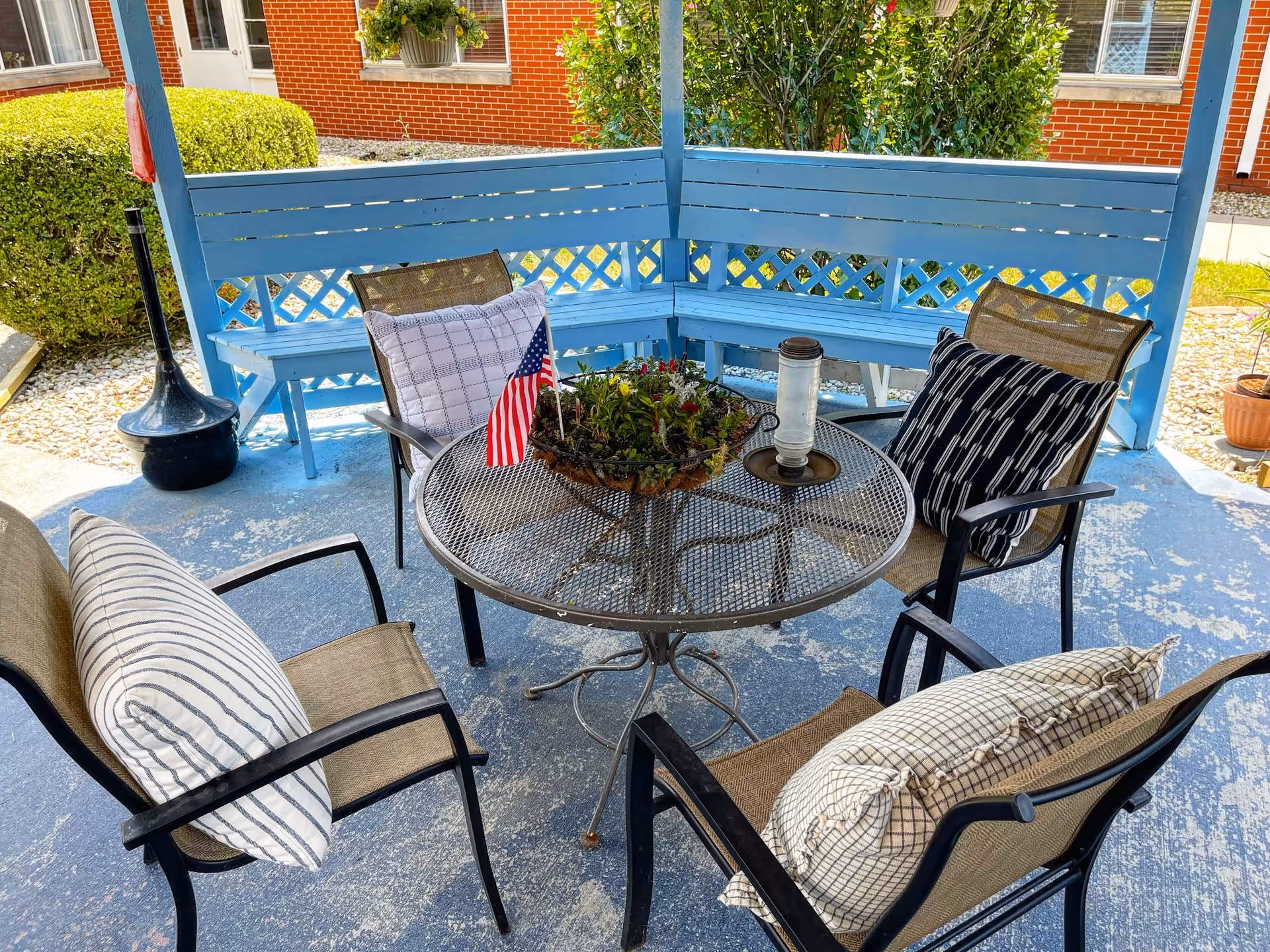 Covered outdoor seating area with a round metal table, four cushioned chairs, a blue bench, potted centerpiece and a small American flag.
