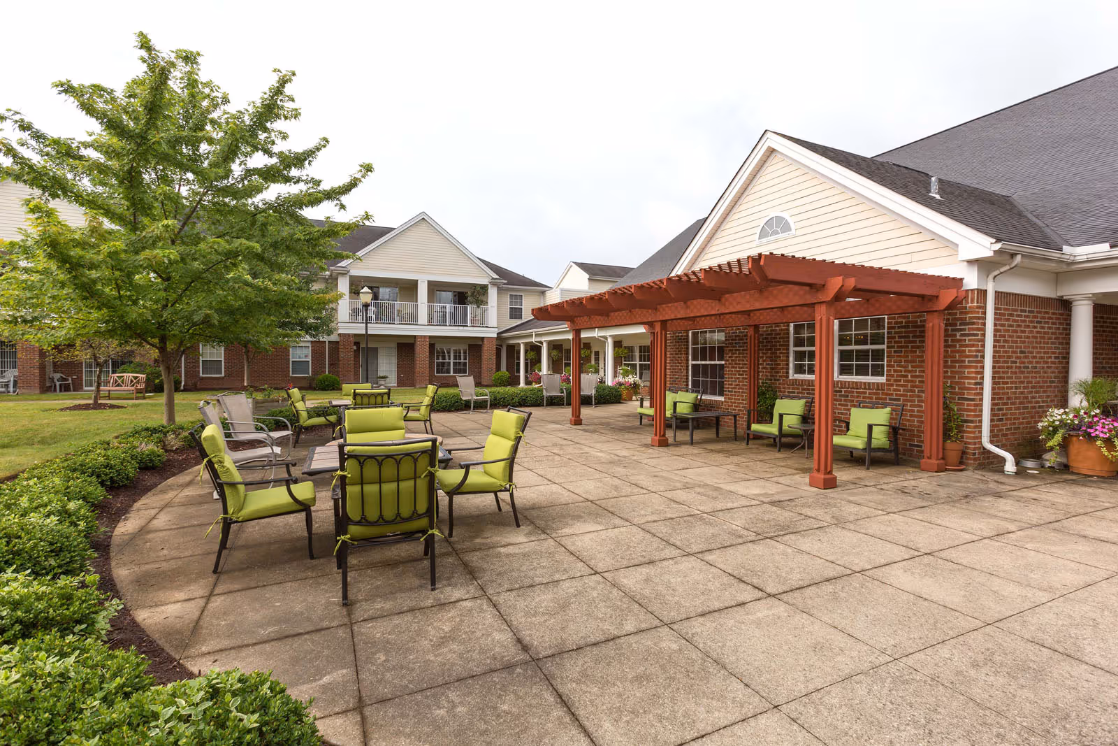 Outdoor patio area at Independence Village of Aurora featuring green cushioned chairs and tables on a paved surface, a wooden pergola attached to a brick building, surrounded by greenery and trees.
