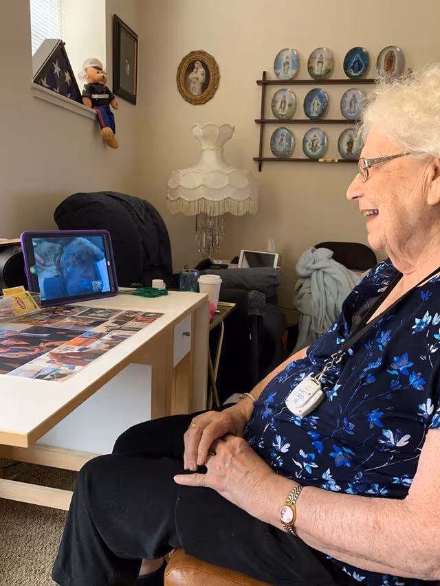 An elderly woman sits in a living room smiling while looking at a tablet on a coffee table showing a video call.