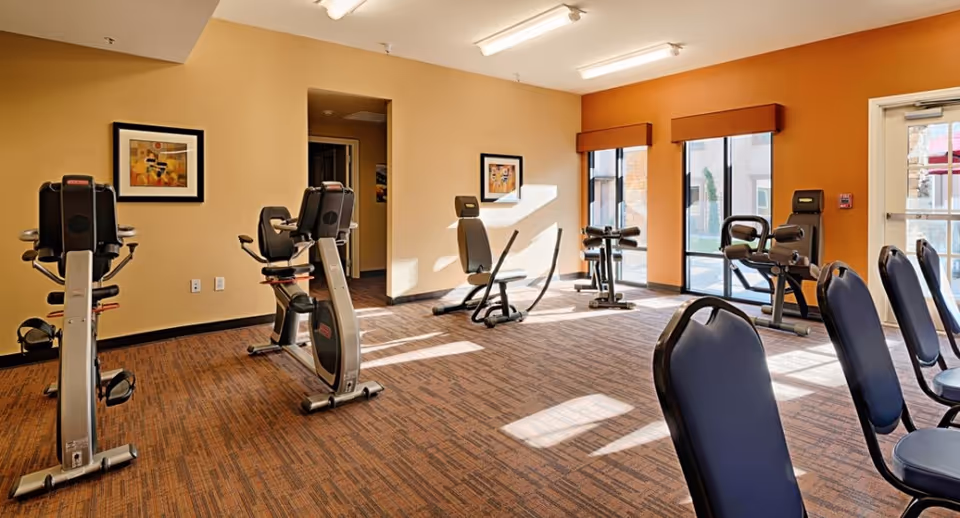 A well-lit exercise room with several stationary exercise bikes and other fitness equipment. The room has orange walls, carpeted flooring, framed artwork on the walls, and large windows letting in natural light. There are also several chairs lined up along one side of the room.