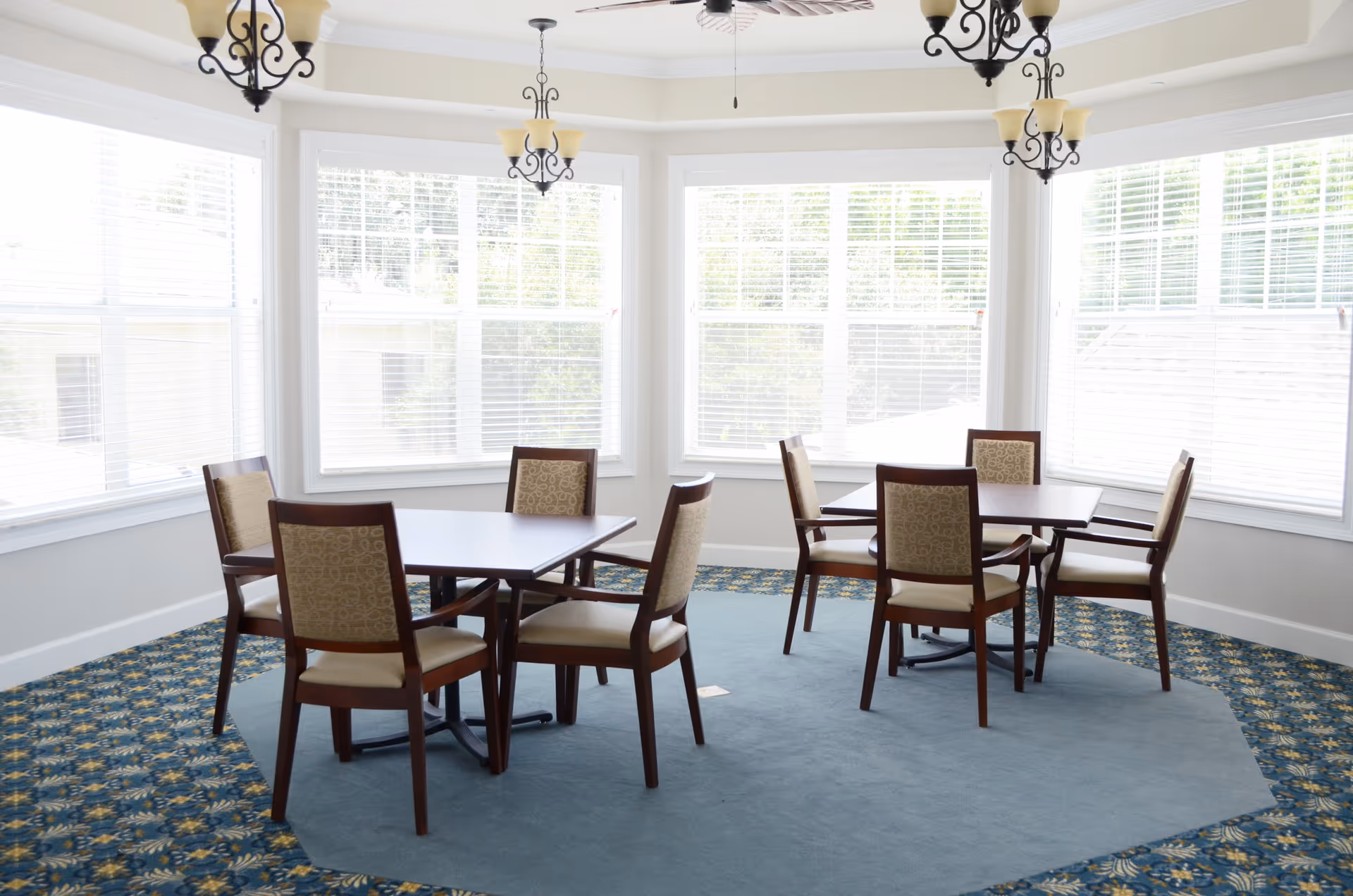A bright dining area with two square wooden tables, each surrounded by four cushioned chairs with patterned backs. The room has large windows with white blinds allowing natural light to fill the space. The floor features a blue carpet with a floral border, and there are three decorative chandeliers hanging from the ceiling.
