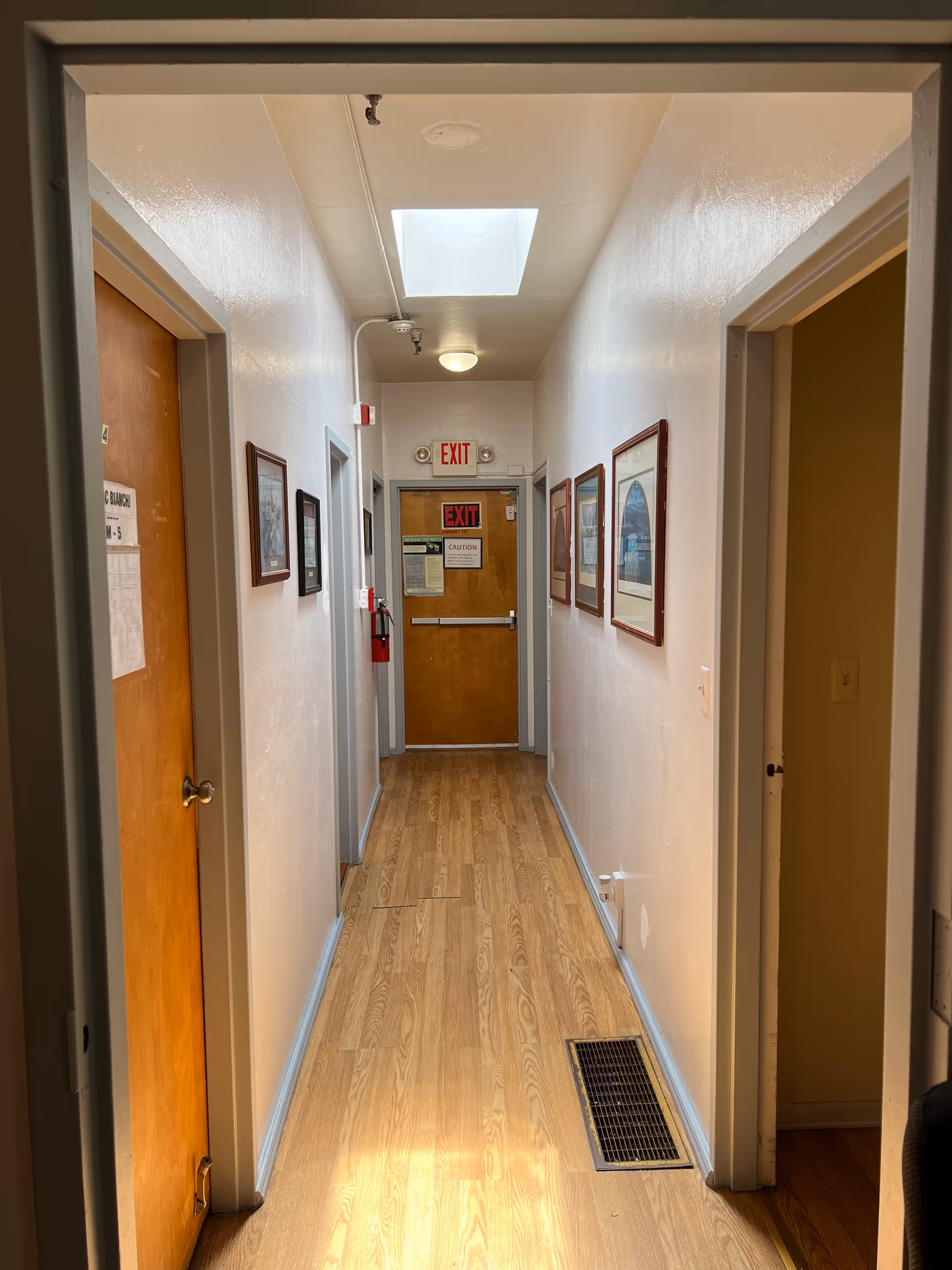 A narrow hallway with light wood flooring and white walls. Several framed pictures hang on the right wall. There are doorways on both sides of the hallway, and an exit door with an illuminated red EXIT sign is visible at the end. A fire extinguisher is mounted on the left wall near the exit door. The ceiling has a skylight and a round light fixture.