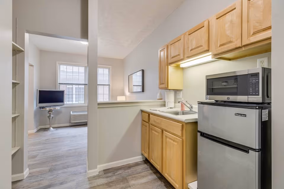 Interior view of a small kitchen area in an assisted living facility with wooden cabinets, a sink, a microwave on top of a mini refrigerator, and a partial view of an adjacent living space with a TV on a stand and two windows.