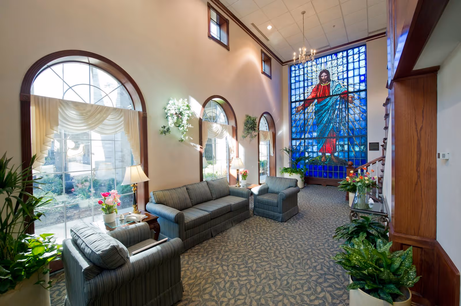 Sunlit common lounge with sofas and armchairs, arched windows, plants, and a tall stained-glass window depicting a robed figure.