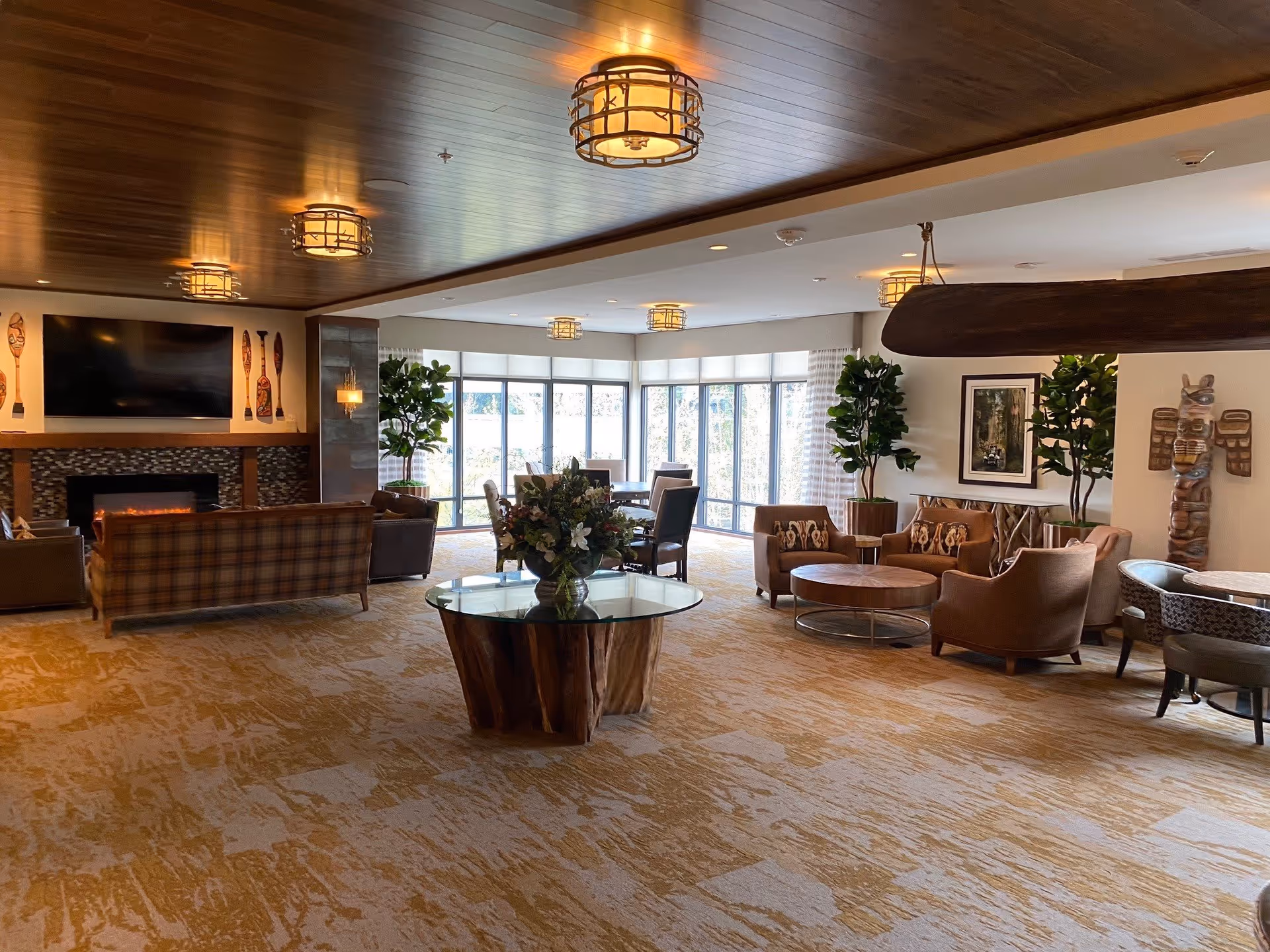 Spacious living room area with a wooden ceiling and warm lighting. The room features a fireplace with a TV mounted above it, several comfortable chairs and sofas arranged around coffee tables, large windows letting in natural light, and decorative plants and artwork including a totem pole on the right side.