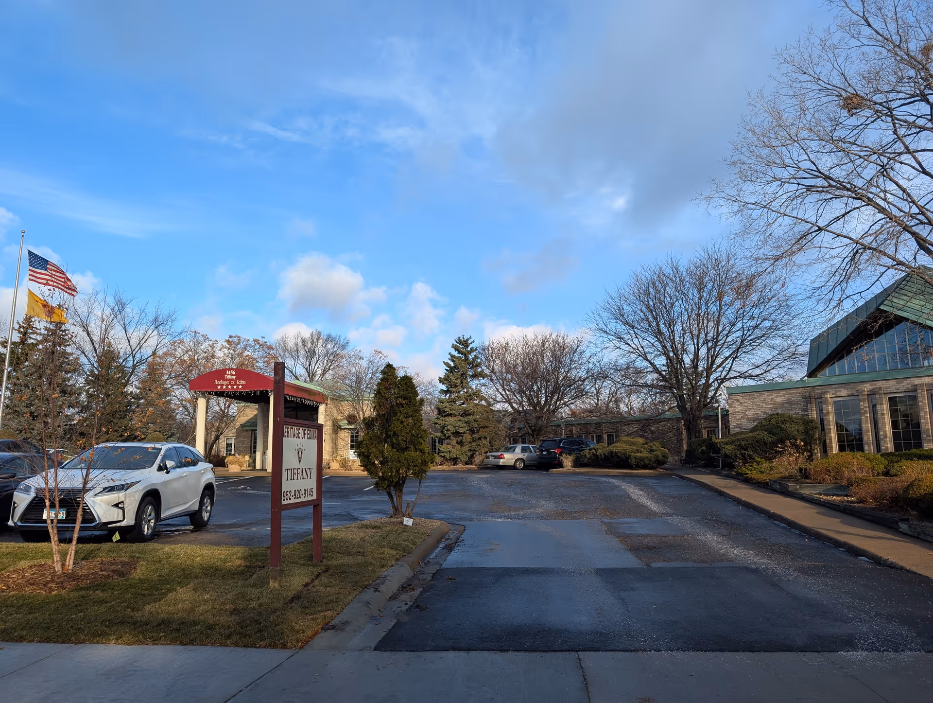 Exterior view of Heritage Of Edina II facility showing a parking lot with several cars, a sign with the facility name and phone number, leafless trees, and a partly cloudy blue sky.