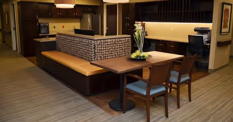 Interior view of a modern common area in a senior living facility featuring a built-in cushioned bench with patterned upholstery, a wooden table with two chairs, a flower arrangement centerpiece, a desk with a computer chair, and cabinetry with under-cabinet lighting.