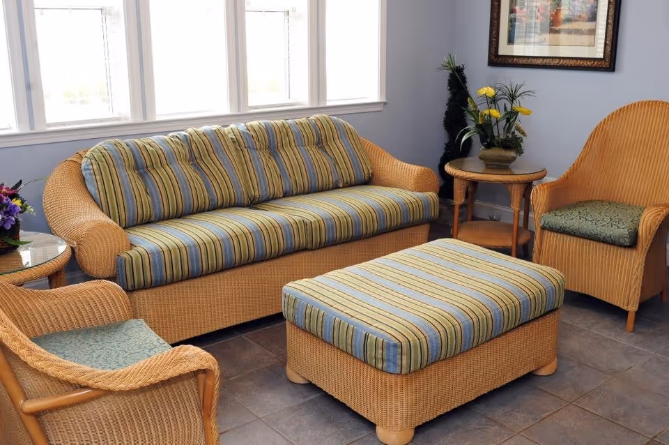 A cozy sitting area with wicker furniture including a striped cushioned sofa, two matching armchairs with green cushions, and a striped cushioned ottoman. There are two small round wicker side tables, one with a flower arrangement and the other with a potted plant. The room has large windows letting in natural light and a framed picture on the wall.